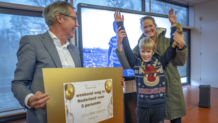 Een man in een pak houdt een grote gouden cheque vast terwijl een vrouw en een kind enthousiast hun armen opsteken.