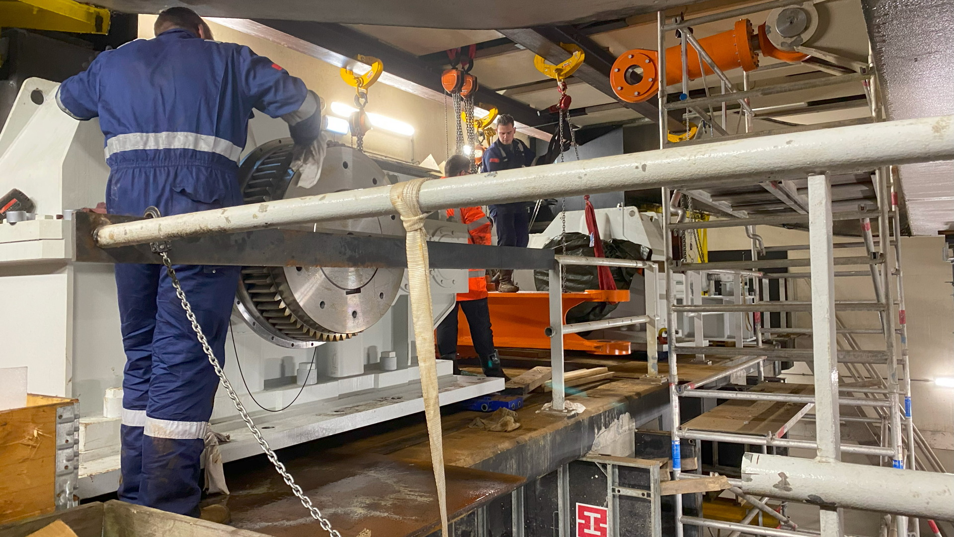 Cluster van drie mensen werkt aan groot tandwielmechanisme op bouwplaats met steiger en hijstools.