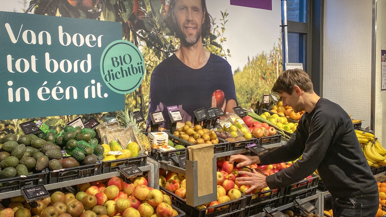 Man kiest fruit bij een supermarktdisplay met de tekst "Van boer tot bord in één rit" en "BIO dichtbij".