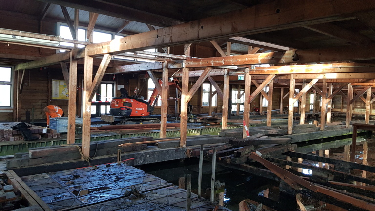 Interieur van een houten gebouw in renovatie, met zichtbare balken en bouwgereedschap, inclusief een kleine graafmachine.