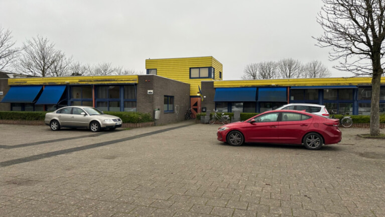 Een laag gebouw met gele en blauwe accenten en meerdere auto's geparkeerd ervoor op een grijze dag.