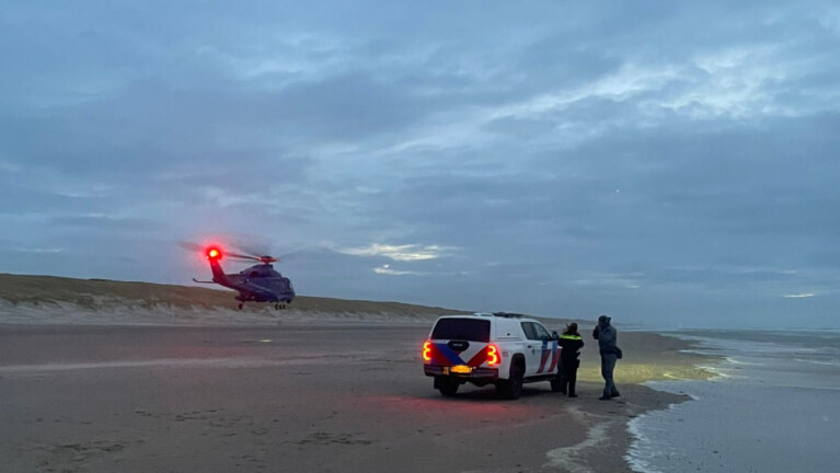 Helikopter landt op een strand bij schemering met een politievoertuig en personen in de buurt.