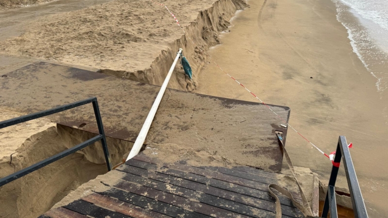 Beschadigde trap en zandduinen aan een kustlijn met rood-wit afzetlint.