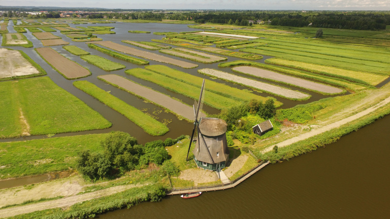 Luchtfoto van een traditionele Nederlandse windmolen omringd door groene weilanden en waterwegen.