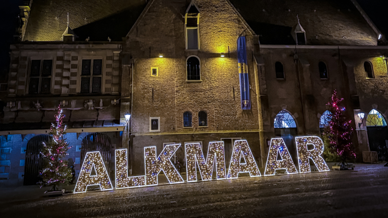 Verlichte letters vormen het woord "ALKMAAR" voor een historisch gebouw, geflankeerd door versierde kerstbomen.