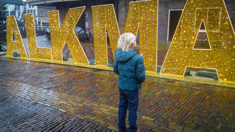 Een kind in een blauwe jas kijkt naar grote, verlichte letters die "ALKMAAR" spellen op een natte, geplaveide straat.