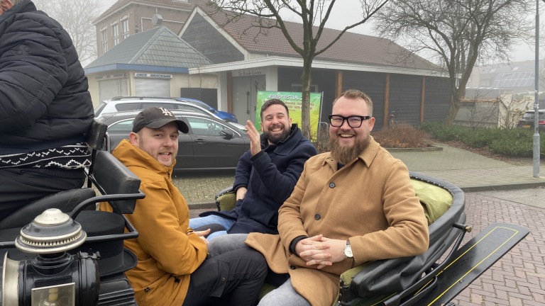 Drie mannen glimlachen in een open koets op een mistige dag in een stedelijke omgeving.