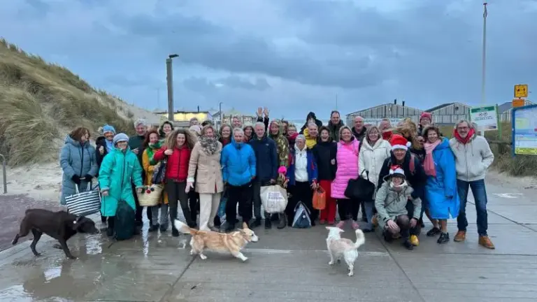 Groep mensen met honden poseren voor een strandduin, sommigen dragen kleurrijke jassen en hoeden.