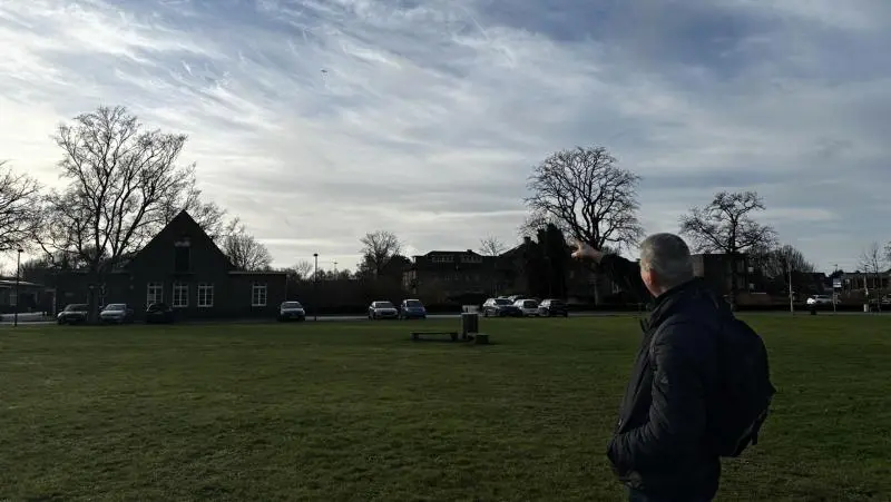 Man op grasveld wijst naar lucht, met bomen en huizen op de achtergrond.