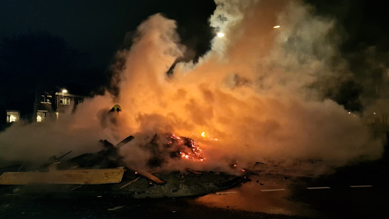 Brandende stapel hout met sterke rookontwikkeling in een nachtelijke straat.