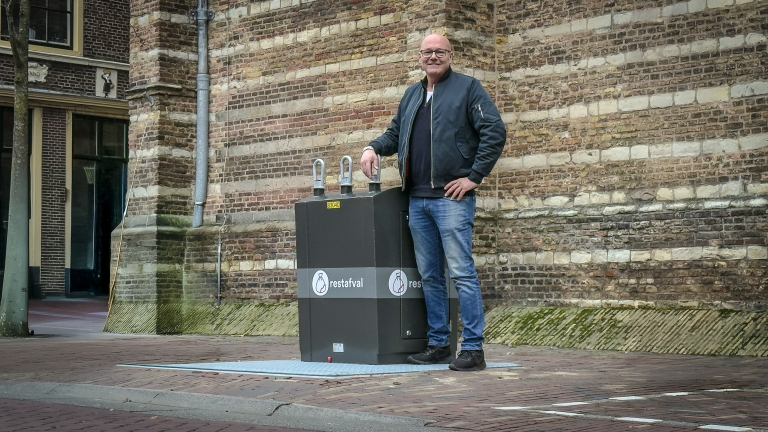 Man poseert naast een ondergrondse vuilcontainer voor restafval tegen een bakstenen gebouw.