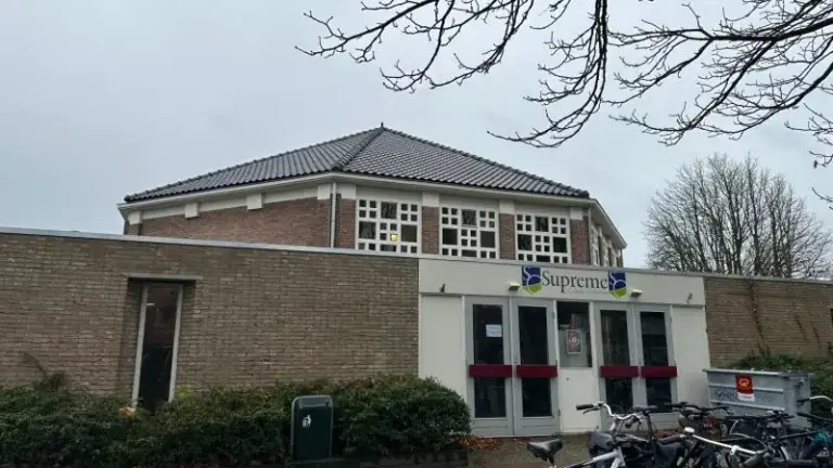 Bakstenen gebouw met grijs dak, ramen en voordeur met een bordje "Supreme", omringd door bomen en een fietsenrek.