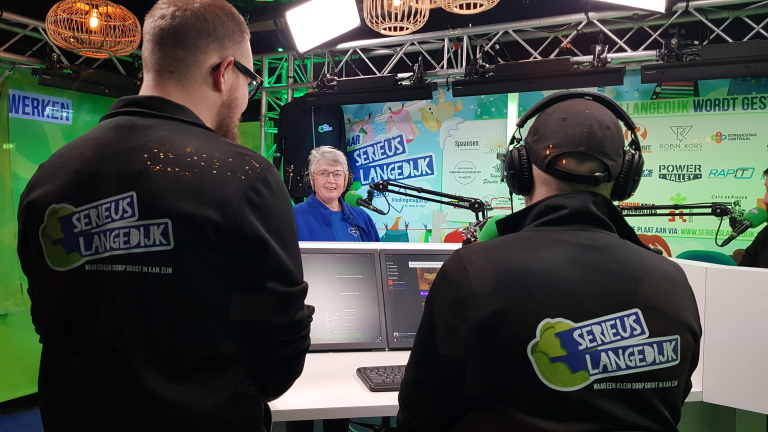 Twee mannen in zwarte jassen met "Serieus Langedijk" op de rug staan tegenover een oudere vrouw met een headset achter een microfoon in een studio.