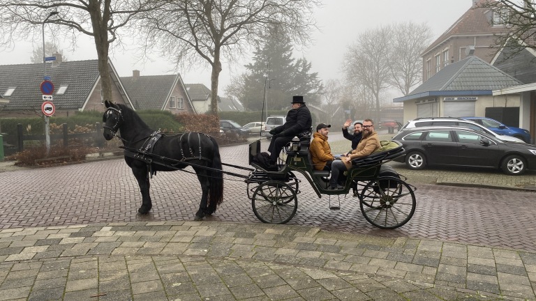 Een koets getrokken door een zwart paard met vier personen, in een mistige straat.