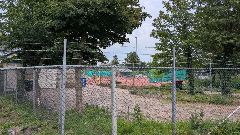 Tennisbanen omgeven door een draadhekken en prikkeldraad, met bomen en een vuilnisbak op de achtergrond.