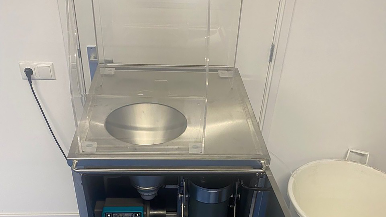Industriële machine met transparante kap en ronde metalen kom op een werkblad, met open onderkast en opvangbak ernaast