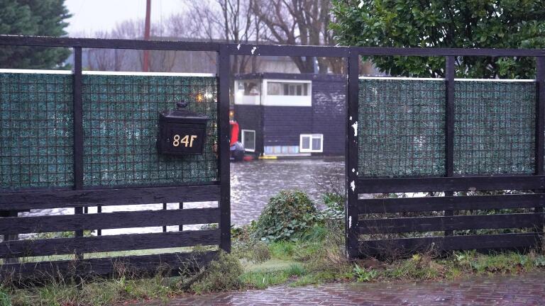 Hek met huisnummer 84F, zicht op een overstroomd gebied met een gebouw op de achtergrond.