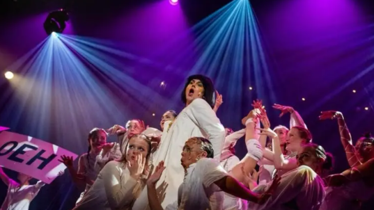 Groep artiesten op een podium met kleurrijke verlichting, een persoon centraal draagt een zwarte hoed en witte kleding, omringd door dansers; iemand houdt een bord vast met het woord "OEH".