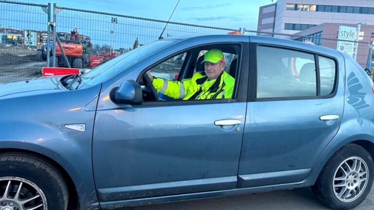 Man in een blauwe auto met reflecterende jas en pet, op een bouwplaats.