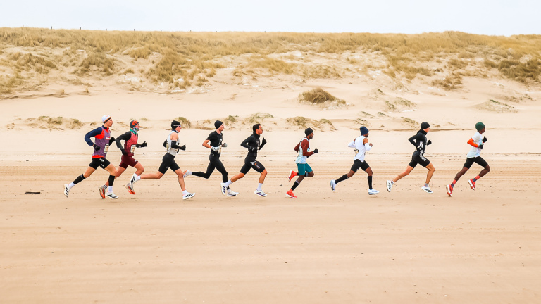 Atleten rennen op een strand met duinen op de achtergrond, gekleed in sportkleding voor koud weer.