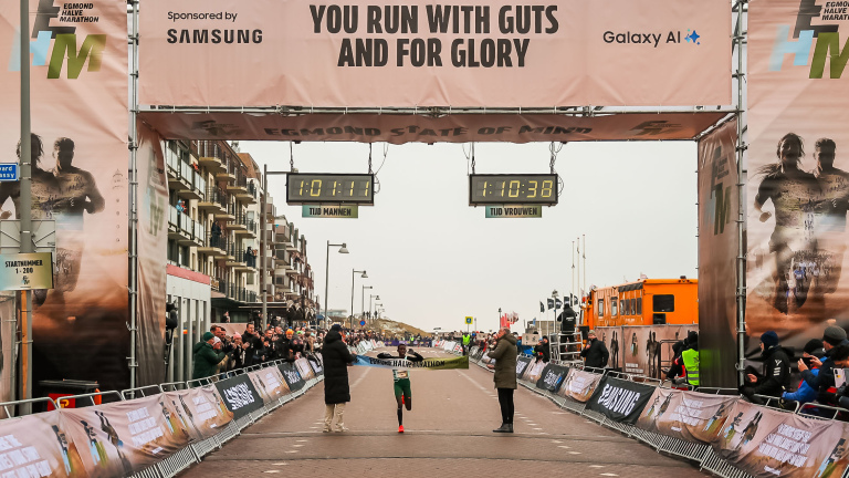Een hardloper passeert de finish van de Egmond Halve Marathon terwijl toeschouwers applaudisseren, met tijdklokken boven.