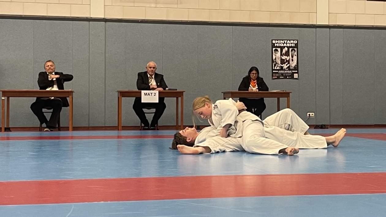 Judo-wedstrijd met deelnemers op de mat, juryleden op de achtergrond en decoratieve Japanse karakters aan de muur.