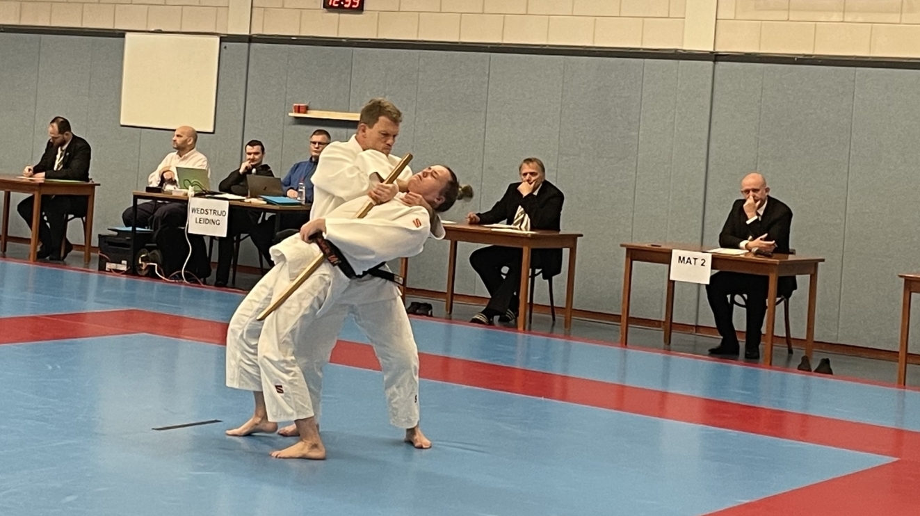 Twee judoka's in actie op een mat binnen een sporthal, met juryleden zittend aan tafels langs de zijkant.