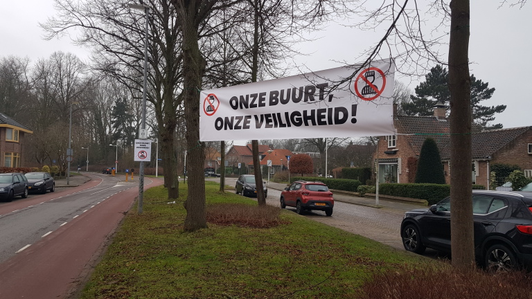 Spandoek aan bomen met de tekst "ONZE BUURT, ONZE VEILIGHEID!" en afbeelding van een gevangenisgebouwen met een verbodsteken. Straat met auto's en huizen op de achtergrond.