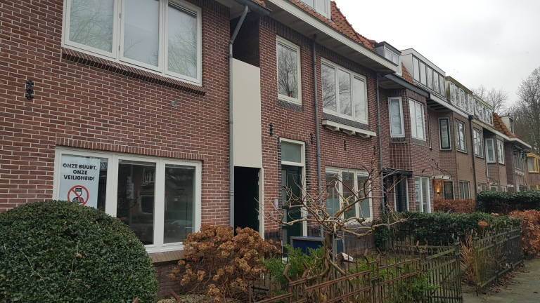 Rij van bakstenen huizen met tuinen; bord met tekst "Onze buurt, onze veiligheid!" en een symbool van een verkeersverbod.