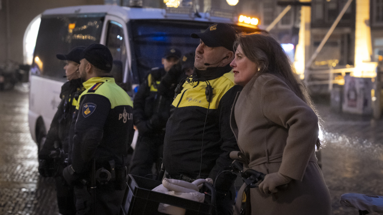 Vrouw in gesprek met politieagenten bij een politiebusje in een nachtelijke stedelijke omgeving.