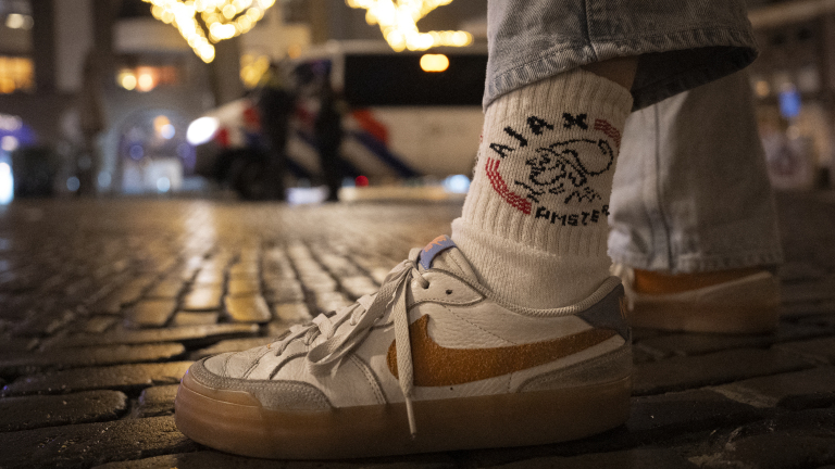 Close-up van een voet met een witte sneaker en een sok met het logo van "Ajax Amsterdam" op een straat met kasseien in een nachtelijke stad. Op de achtergrond zijn lichtjes en een vaag politiemotief zichtbaar.