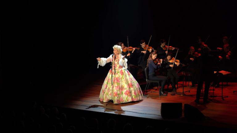 Zangeres in bloemrijke jurk zingt op podium, begeleid door orkest.