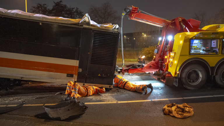 Twee personen in oranje werkkleding werken 's nachts met een takelwagen om een gekantelde vrachtwagen rechtop te krijgen op een weg.