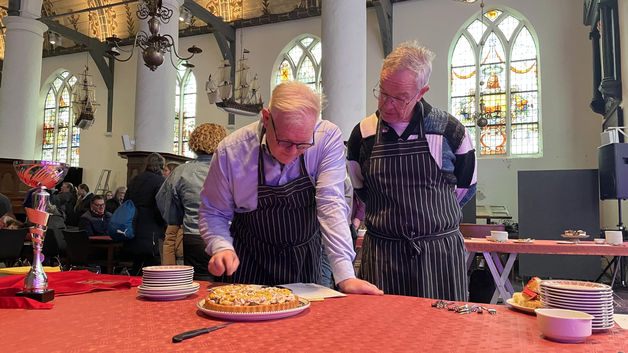 Twee mannen in schorten staan voor een taart op een rode tafel in een kerk, met een trofee en stapels borden.