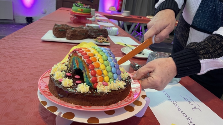 Een persoon snijdt een regenboogkleurige taart aan op een gedekte tafel.