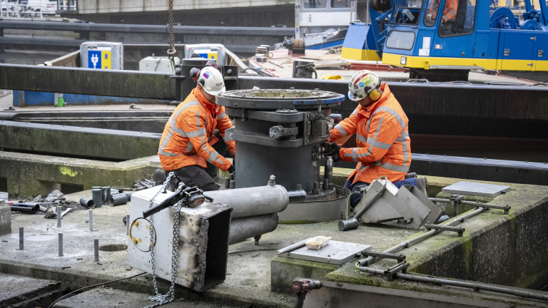 Twee bouwvakkers in oranje veiligheidskleding werken aan zware machines op een bouwplaats met een blauwe kraan op de achtergrond.