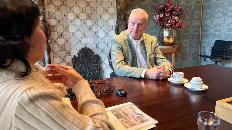 Twee personen zitten aan een tafel in een stijlvolle kamer met gedecoreerde muren; er staan kopjes en een boek op tafel.