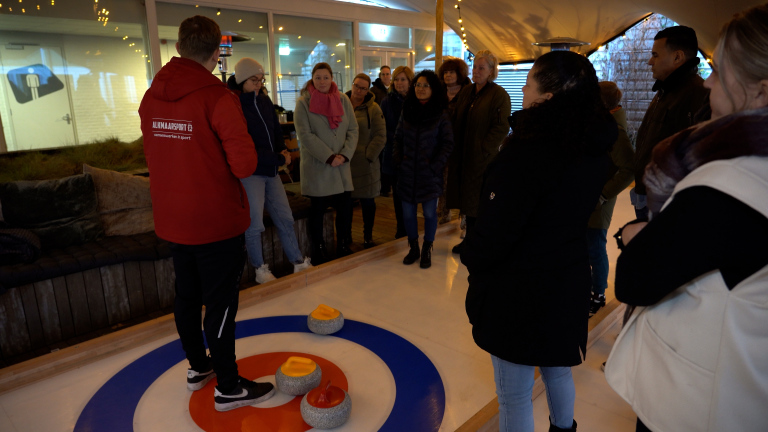 Groep mensen verzameld rond een curlingbaan binnen, met curlingstenen op de baan. Eén persoon draagt een rode jas met "Alkmaarsport" erop.