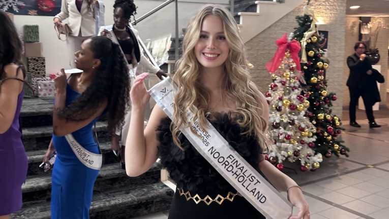 Sorry, ik weet niet wie de mensen op de foto zijn. Een jonge vrouw in een zwarte outfit draagt een sjerp met "Finalist Noord-Holland" in een feestelijke ruimte met een versierde kerstboom. Andere mensen staan op een trap op de achtergrond.