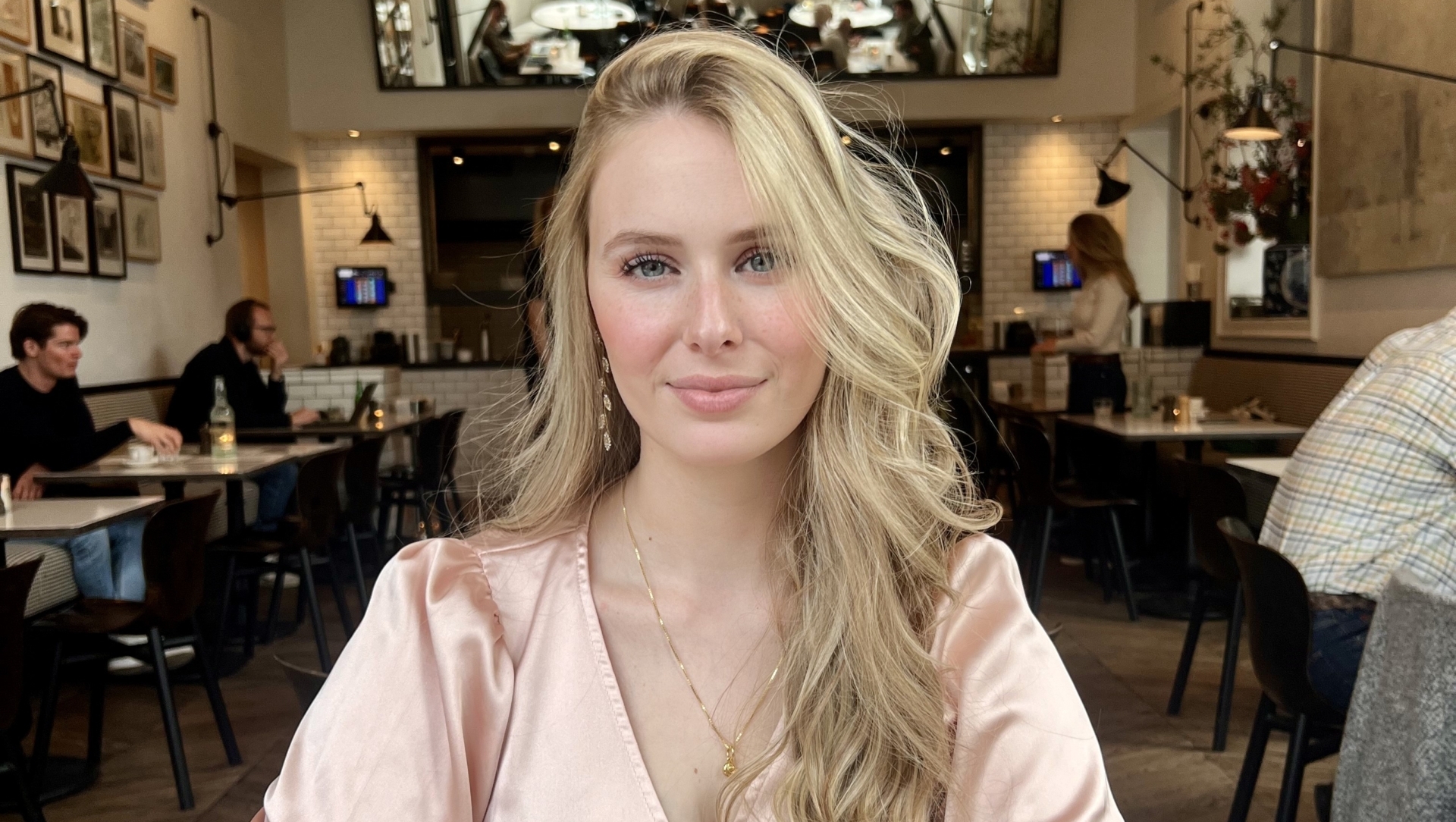 Een vrouw met blond haar zit aan een tafel in een café met een glas groene thee voor zich.