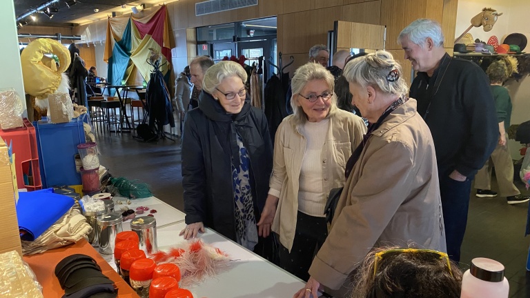 Drie oudere vrouwen praten met elkaar in een kamer vol met kleding en rekwisieten. Op de tafel liggen verschillende materialen en accessoires. In de achtergrond staan andere mensen te praten.