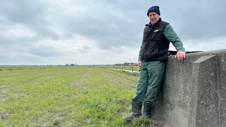 Man in werkkleding leunt tegen een betonnen muur in een weids veld onder een bewolkte lucht.