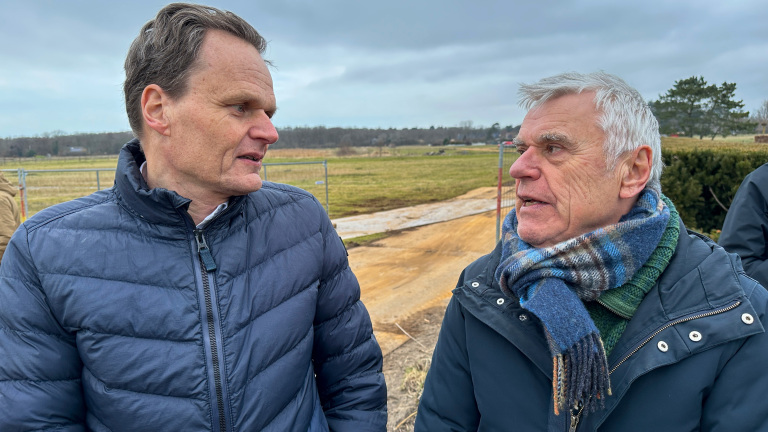 Twee mannen in winterjassen converseren buiten op een grasveld met een bewolkte lucht.