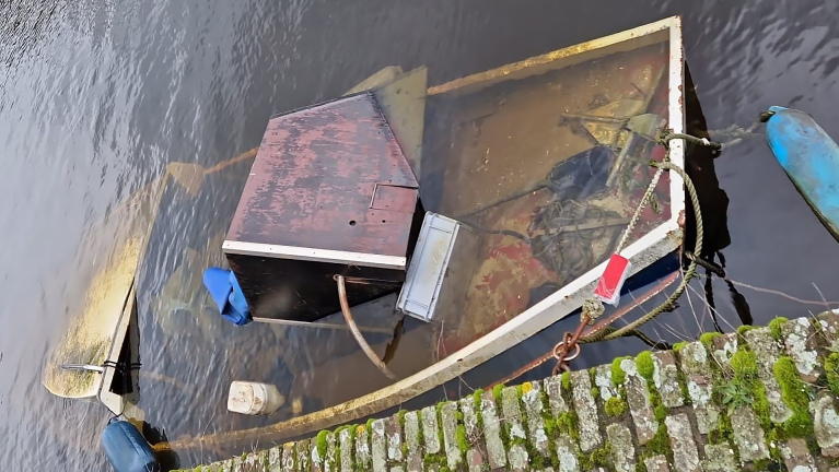 Een half gezonken boot in een waterweg, met een met mos begroeid stenen pad op de voorgrond.