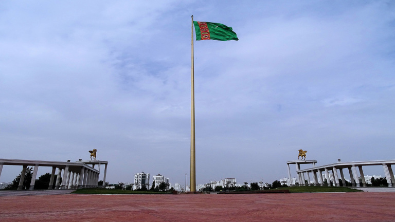 Grote vlaggenmast met de vlag van Turkmenistan op een plein, omgeven door pilaren en gouden standbeelden, met stad op de achtergrond.