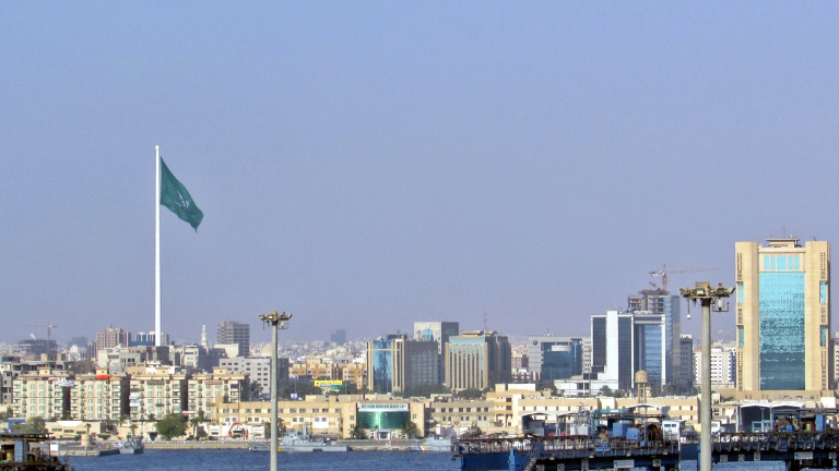 Panorama van een stad met een grote vlag van Saudi-Arabië op een hoge vlaggenmast, omringd door moderne gebouwen en een waterkant op de voorgrond.