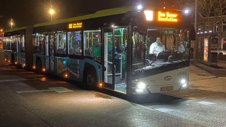 Een gelede bus van vervoerder "Overal" met bestemming "Den Oever Busstation" stopt ‘s avonds bij een halte.