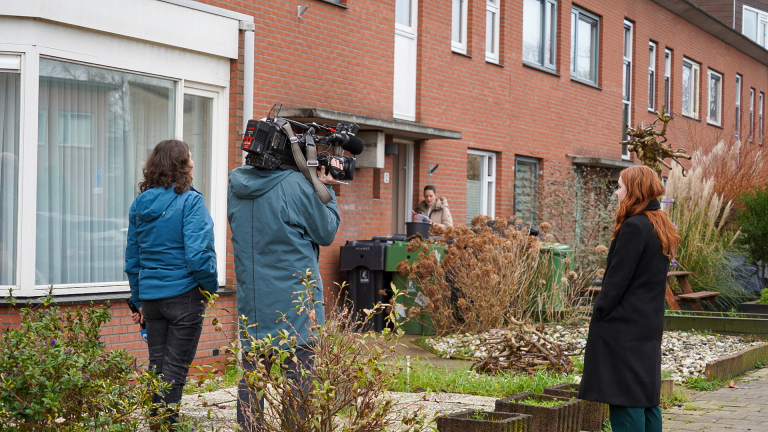 Een cameraploeg filmt een vrouw bij de ingang van een bakstenen huis.