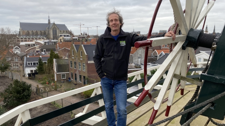 Een man staat op een molenplatform met een stad op de achtergrond, waaronder een grote kerk.