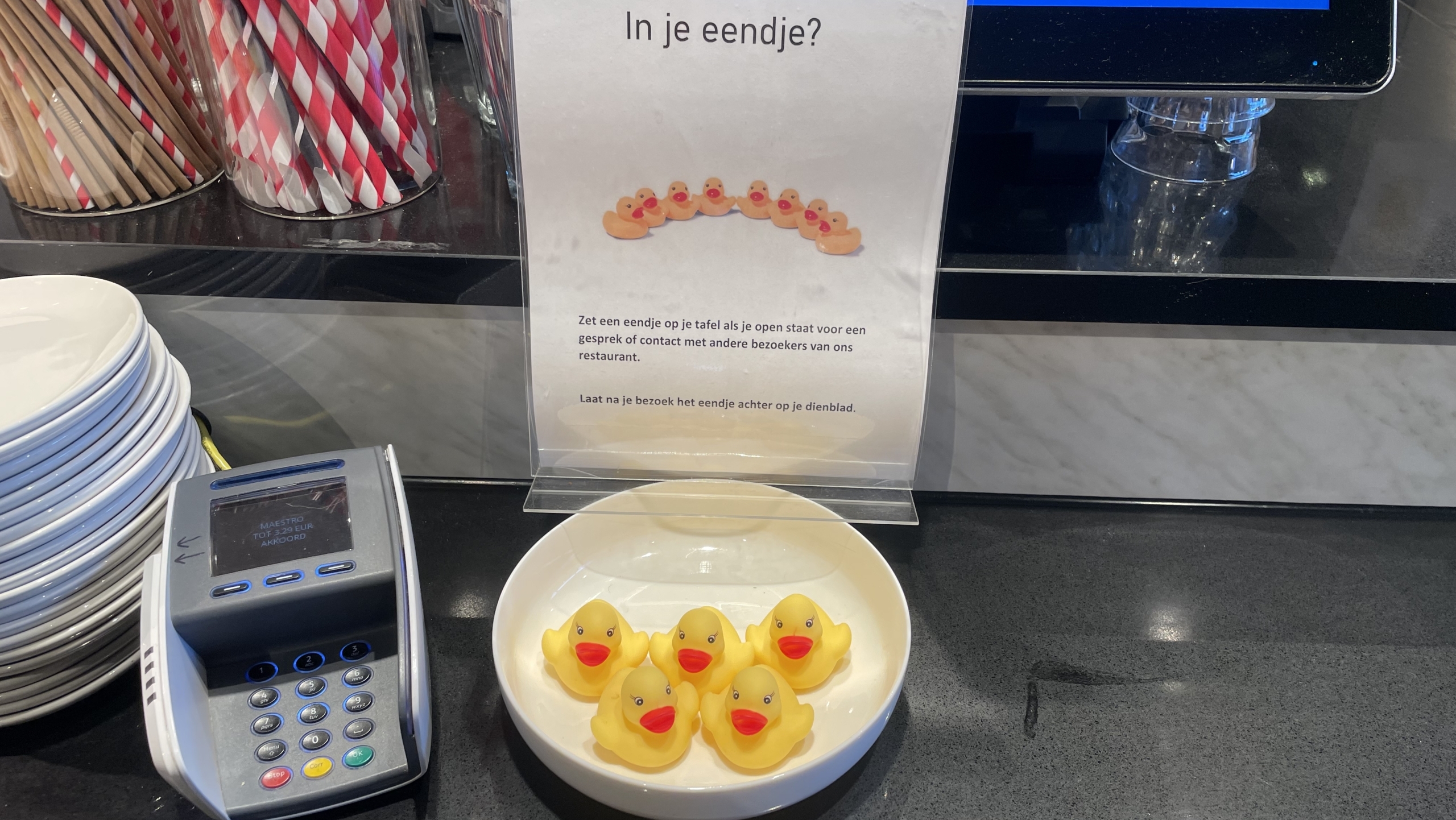 Betalingsautomaat naast een kom met gele badeendjes en een bord met instructies in een restaurant.